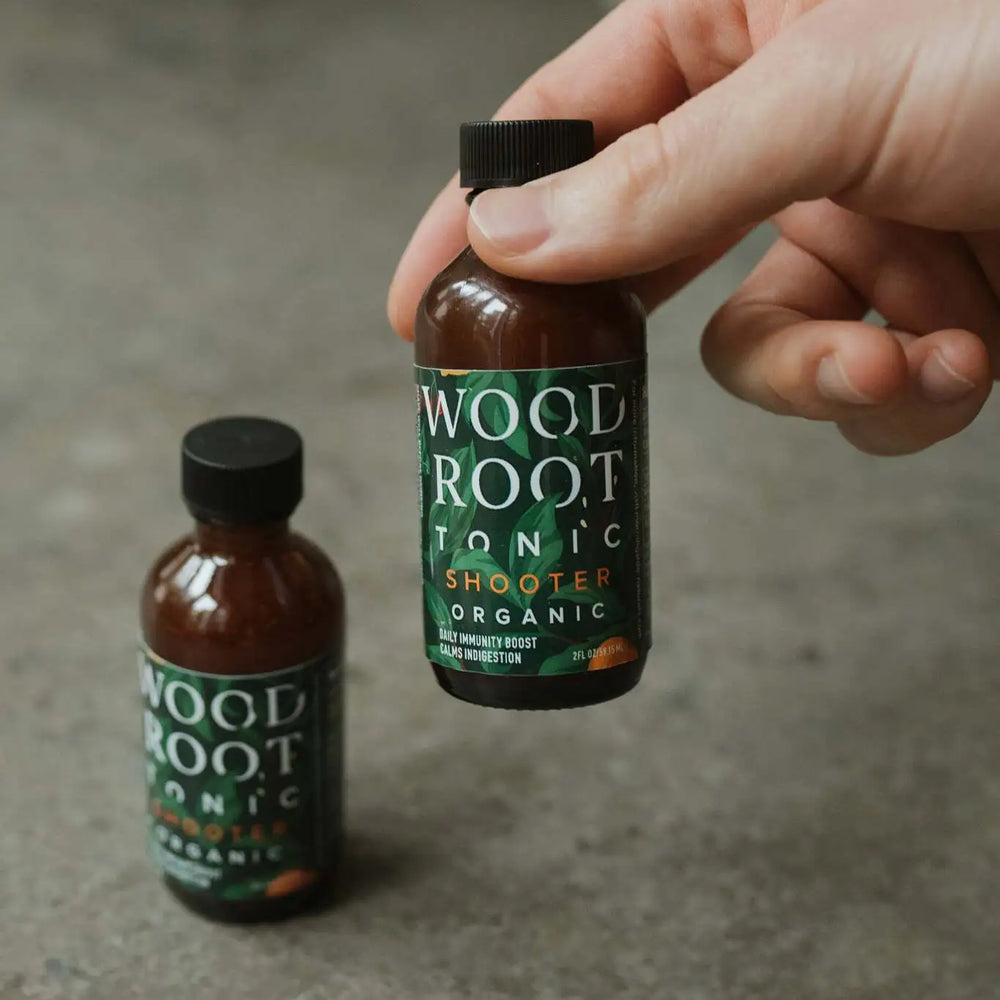 Hand picking up Woodroot Tonic shooter bottle, with a shooter in the background.