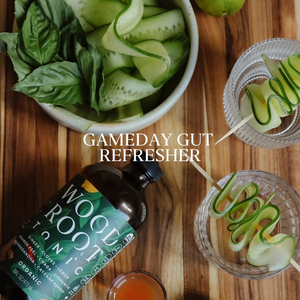 Woodroot tonic bottle on a wooden cutting board being used to make a gut healthy cocktail with cucumber and basil.