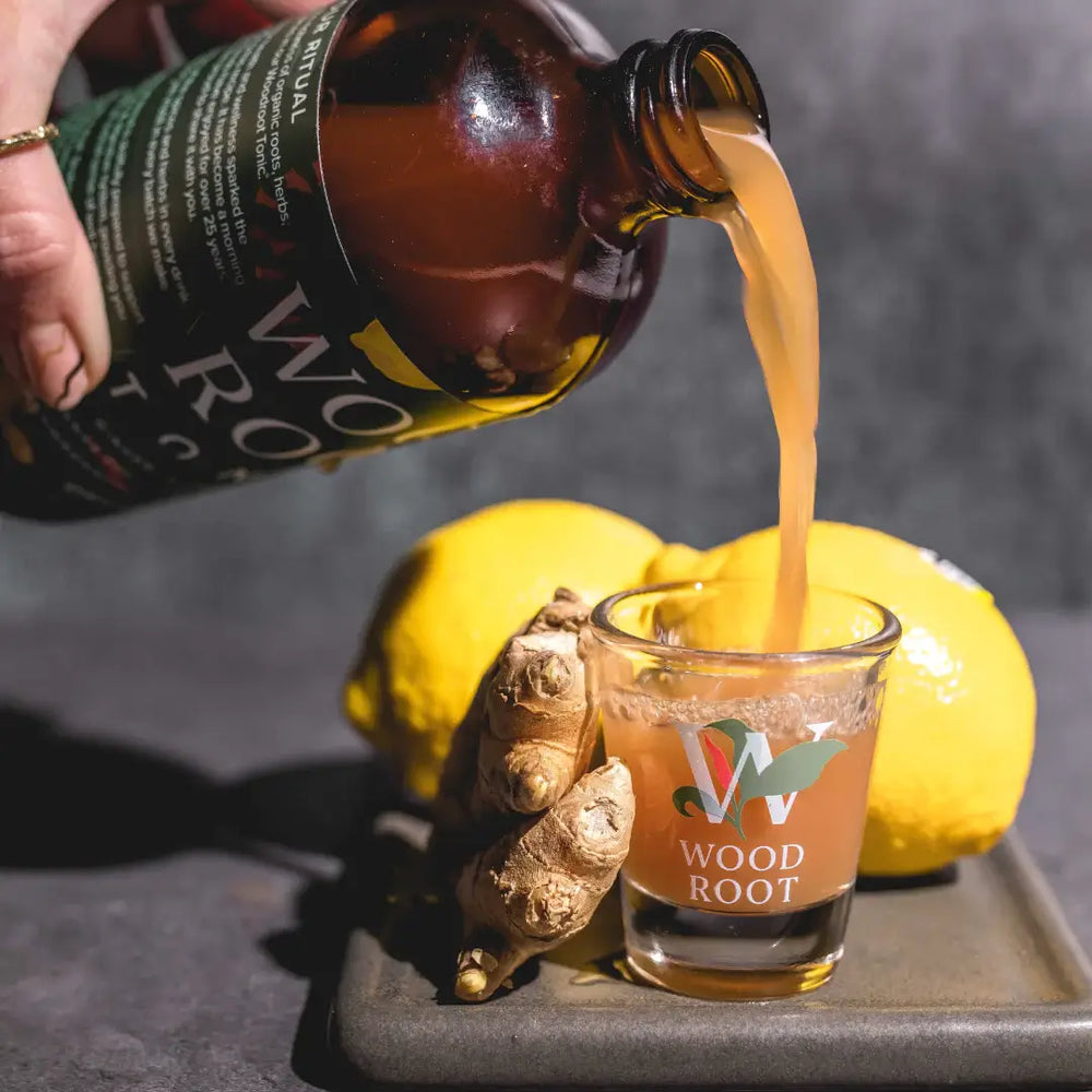 
                  
                    hand pouring organic woodroot tonic gut shot into shot glass surrounded by lemon and ginger. 
                  
                