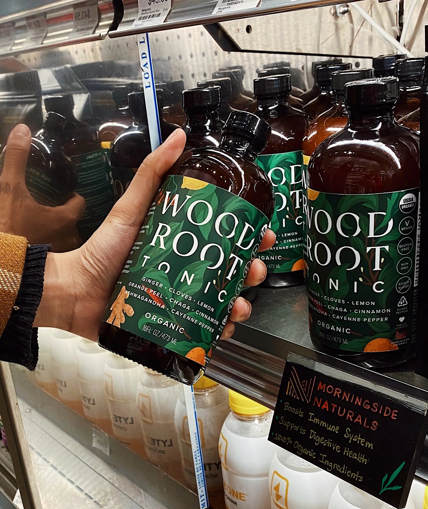 Hand picking up Woodroot Tonic bottle from the shelves of a store. 