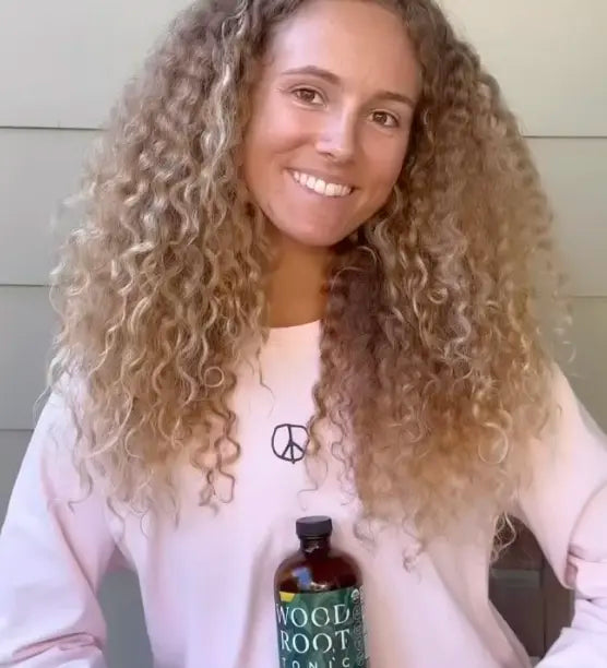 
                      
                        Woman smiling holding Woodroot Tonic herbal gut shot. 
                      
                    