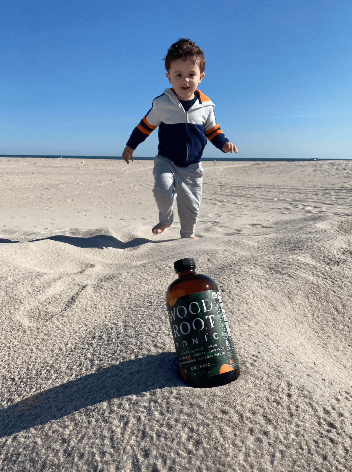 Little boy running towards bottle of Woodroot tonic at the beach.