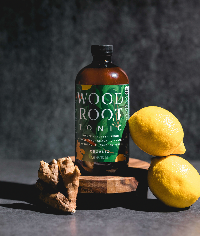 USDA Organic Woodroot Tonic bottle with ginger and lemons, for gut health and inflammation relief, on a wooden stand against a grey backdrop.
