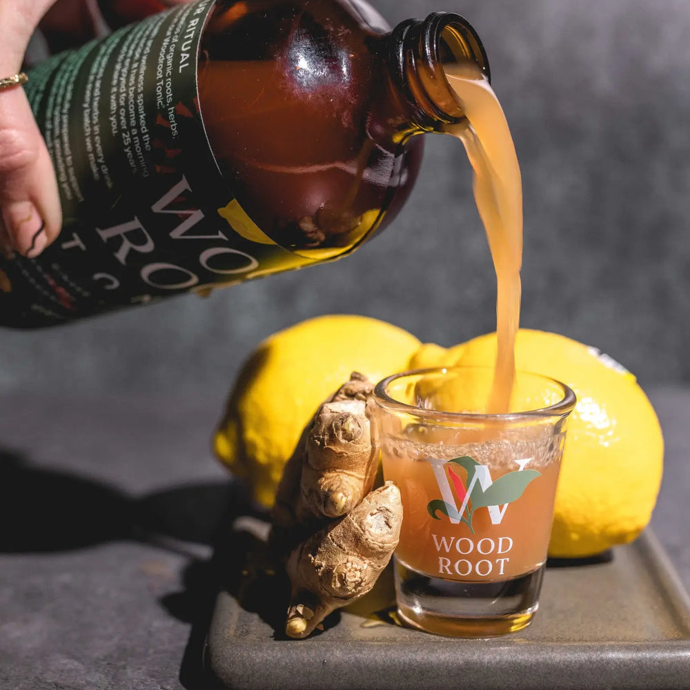 Hand pouring Woodroot Tonic into shot glass, surrounded by anti-inflammatory ingredients ginger and lemon. 