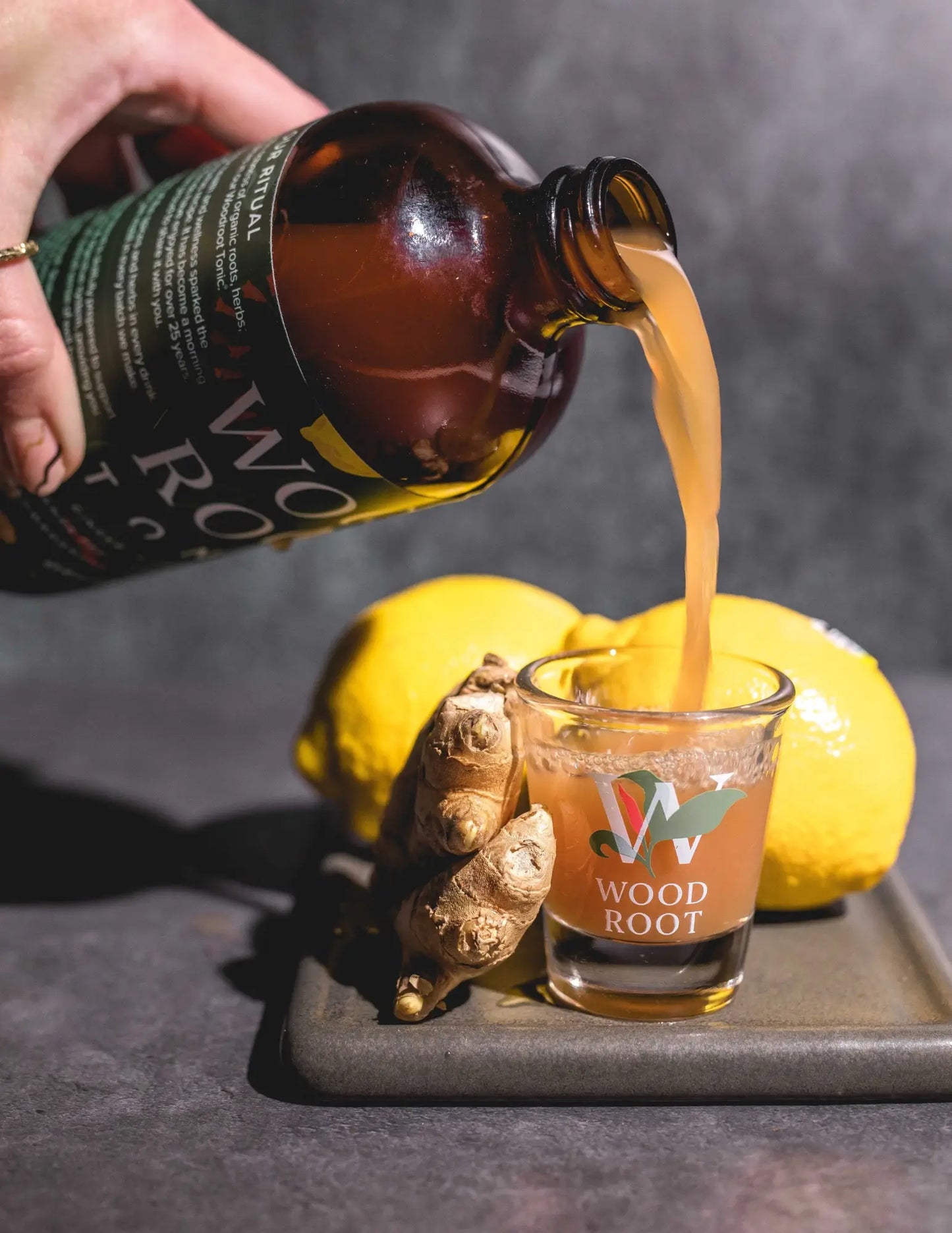 Hand pouring Woodroot Tonic into shot glass, surrounded by anti-inflammatory ingredients ginger and lemon. 