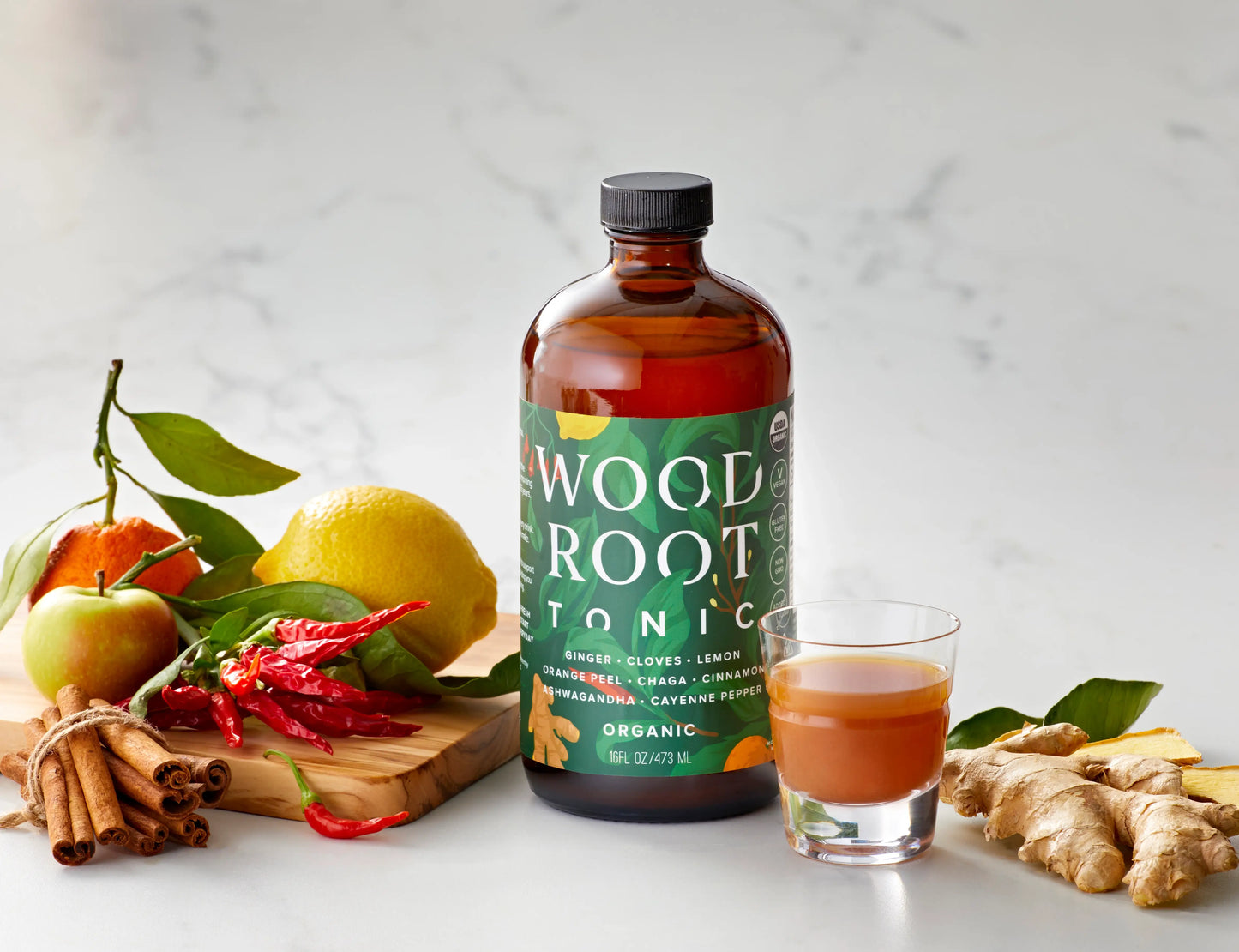 Woodroot Tonic bottle with gut shot already poured in glass shooter, surrounded by usda organic ingredients. 