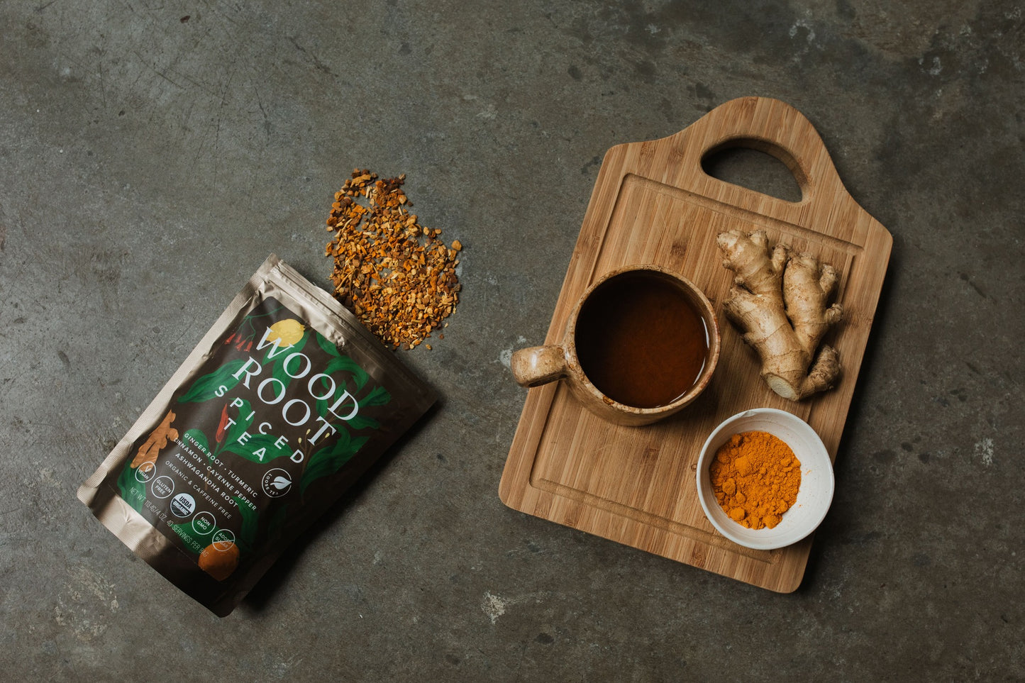 "A bag of Woodroot Spiced Tea with its contents spilled out beside a wooden cutting board, which holds a cup of brewed tea, fresh ginger, and a bowl of turmeric powder.