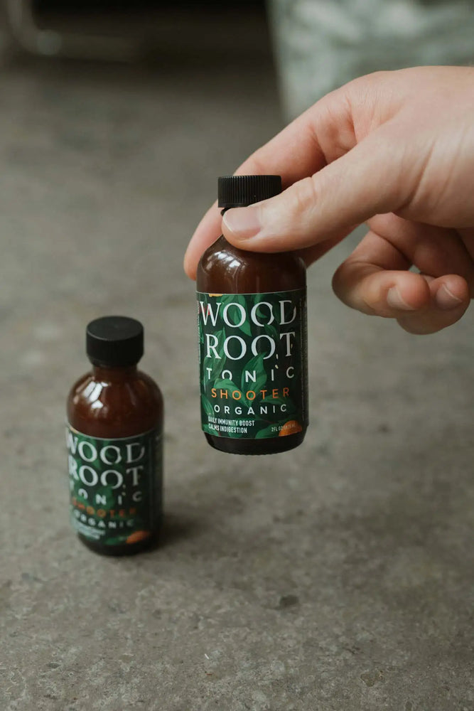 Hand picking up Woodroot Tonic shooter bottle, with a shooter in the background.