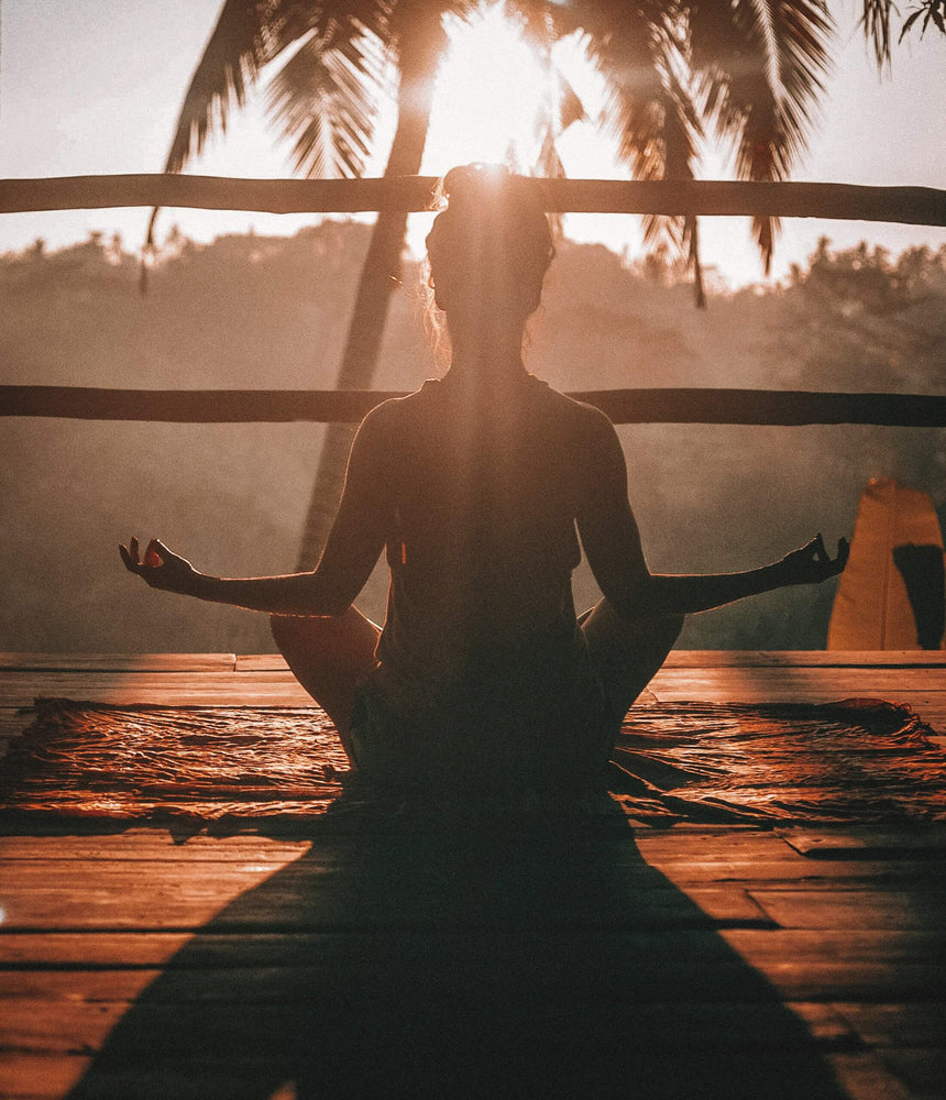woman meditating in the morning on a wood deck with a palm tree in front of her.